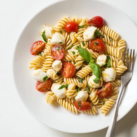 A close-up of Caprese Salad Pasta shows glossy olive oil drizzled over fusilli, tomatoes, and mozzarella, garnished with torn basil leaves.