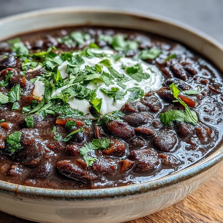 A ladle pours rich Black Bean Soup topped with sour cream and lime wedges.